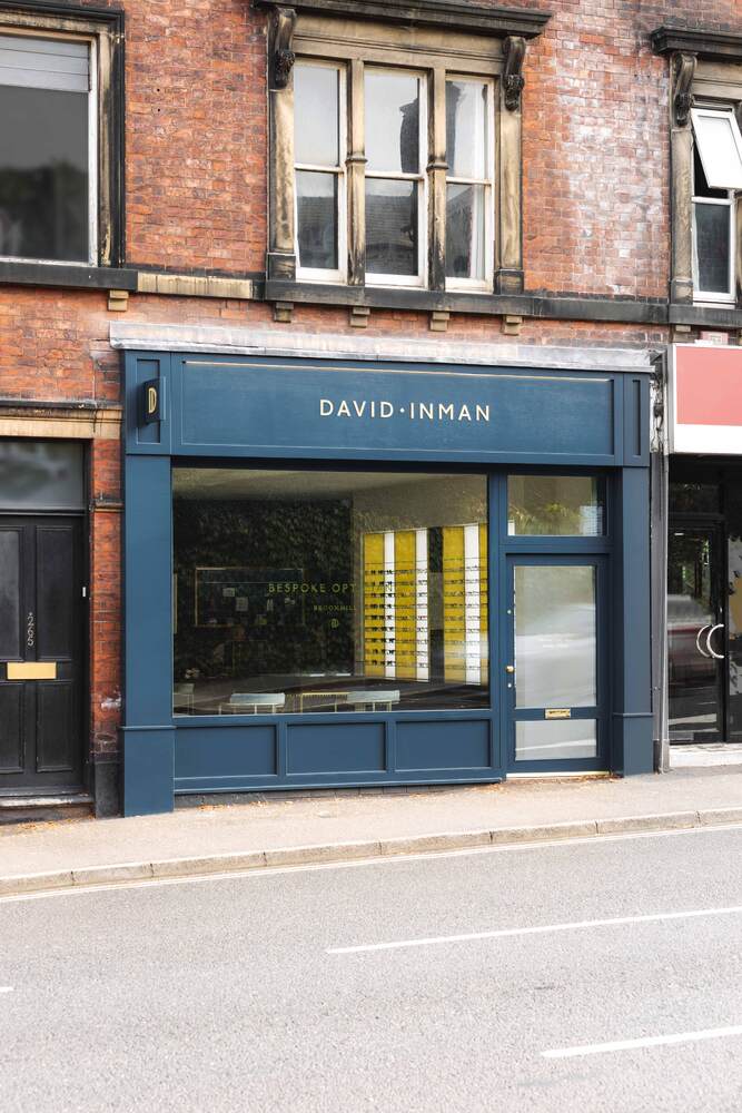 David Inman Bespoke Opticians dark blue shop front in red brick building. Yellow and white display inside holding glasses.